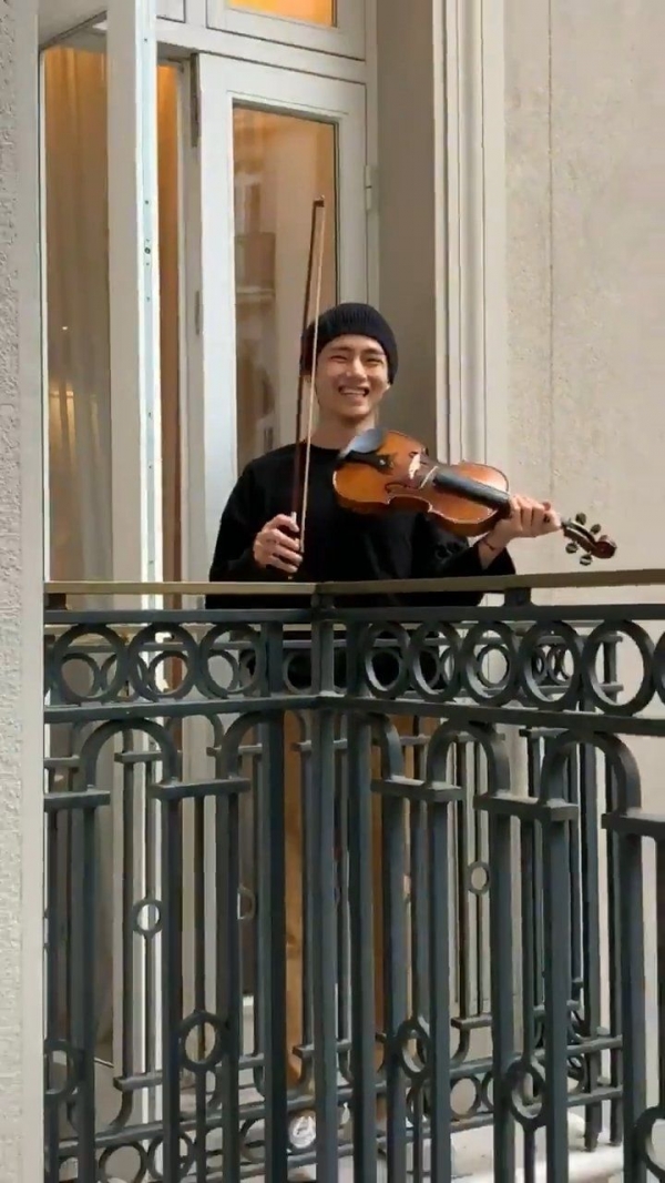 방탄소년단의 뷔가 난간에서 바이올린을 켜고 수줍은 미소를 짓고 있다.